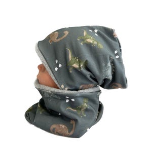 Può includere: Un set di cappello e scaldacollo grigio con motivo a dinosauri. Il tessuto presenta dinosauri verdi e marroni e impronte bianche. L'interno del cappello e dello scaldacollo è foderato con un morbido materiale grigio.