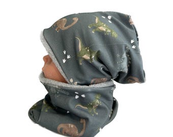 May include: A gray dinosaur-themed beanie hat and neck warmer set. The fabric features a pattern of green and brown dinosaurs and white footprints. The interior of the hat and neck warmer is lined with a soft, gray material.