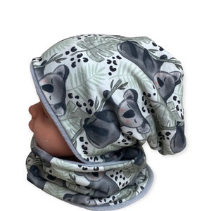 Puede incluir: Un gorro y un cuello de punto a juego de color gris y blanco con estampado de koala. El estampado presenta koalas durmiendo en ramas con hojas verdes y manchas negras.