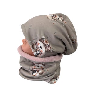 Puede incluir: Conjunto de gorro y braga de cuello grises con un estampado repetido de un oso sonriente con sombrero floral y un pingüino. La braga tiene forro polar rosa. El conjunto es para niños.