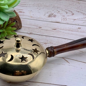 May include: A brass incense burner with a wooden handle. The burner has a round shape with star and crescent moon cutouts. A small brass knob sits atop the lid. The handle is dark brown.