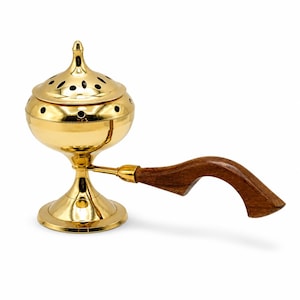 May include: A gold-coloured incense burner with a wooden handle. The burner has a round base, a bulbous body, and a lid with small holes. The handle is curved and made of dark brown wood. The incense burner is designed to hold and burn incense.