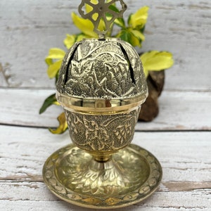 May include: A gold metal incense burner with an ornate design. The burner has a round base and a dome-shaped top with a cross-shaped finial. The burner is sitting on a white wooden surface.