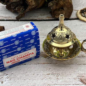 May include: A blue box of Satya Sai Baba Nag Champa Dhoop Cones next to a brass incense burner with a handle. The burner has an ornate design and is sitting on a wooden surface.