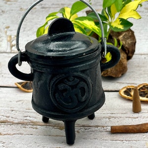 May include: Black cast iron cauldron with a lid and a handle. The cauldron has three legs and an Om symbol on the front. The cauldron is sitting on a wooden surface.