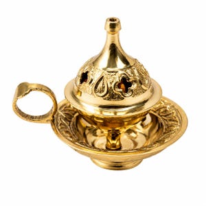 May include: A gold-coloured incense burner with a round base, a decorative plate, and a domed lid with floral cutouts. The burner has a small handle and a tall, slender top.