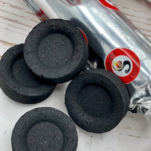 May include: Four black charcoal discs for shisha or hookah, with a silver foil package in the background. The package has the text "Sunlight Charcoal" and a red logo with a flame and the letter "S".