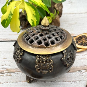 May include: A black and gold metal incense burner with intricate designs and a decorative lid. The burner has three legs and a lattice-like top.