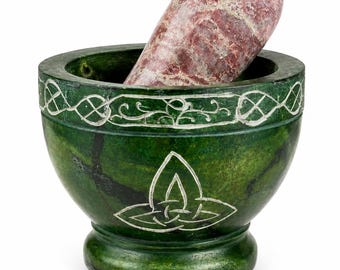 May include: A green mortar and pestle set. The mortar is a deep green with white Celtic knot designs. The pestle is a reddish-pink stone. The mortar has a wide rim and a rounded base. The pestle is oval-shaped.