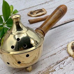 May include: A brass incense burner with a wooden handle. The burner has a round, domed top with holes for incense to burn through. The handle is attached to the side of the burner and is made of dark brown wood.
