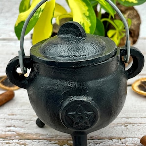 Peut inclure: Un petit chaudron en fonte noire avec un pentagramme sur le devant. Le chaudron a un couvercle et une poignée. Il est posé sur une surface en bois.