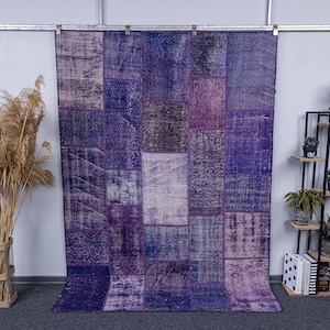 Peut inclure: Un grand tapis rectangulaire en patchwork dans les tons de violet, de lavande et de bleu. Le tapis est composé de divers patchs rectangulaires avec différentes textures et motifs. Le tapis est suspendu à une barre métallique contre un mur gris clair.