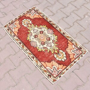May include: A rectangular, hand-woven rug with a central medallion design. The rug features a red field with cream, yellow, and orange floral motifs. The border is a cream colour with a repeating pattern. The rug is approximately 60 x 90 cm.
