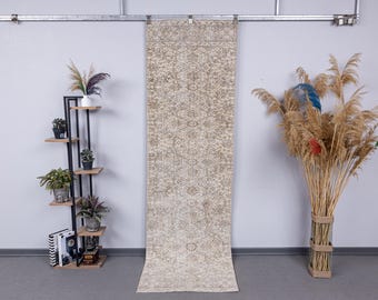 May include: A beige and cream-coloured runner rug with an intricate floral pattern. The rug is displayed vertically against a light grey wall. A black metal plant stand with wooden shelves and potted plants is on the left.