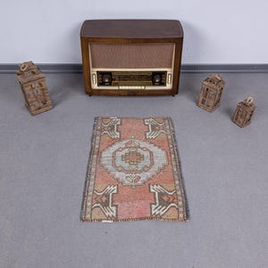 May include: A small, rectangular rug with a central medallion design in shades of pink, gray, and beige. The rug is surrounded by a border with geometric patterns. A vintage radio and several decorative wooden lanterns are also in the image.
