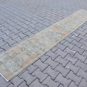 May include: A long, narrow rug with a beige background and a light green pattern. The rug has a distressed look and is laid out on a grey brick surface.