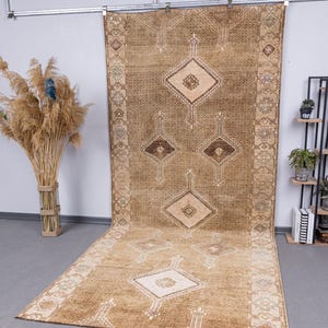 Può includere: Un grande tappeto rettangolare marrone con un motivo geometrico a rombi. Il tappeto presenta un disegno centrale di forme a rombi ripetute in varie tonalità di marrone e beige. Il bordo presenta un motivo dettagliato e ripetitivo. Il tappeto è esposto su un pavimento grigio.