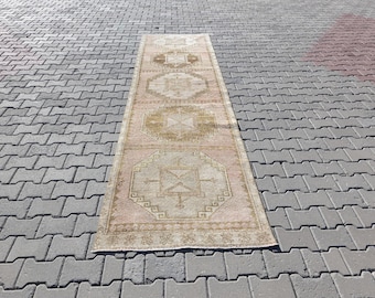 Puede incluir: Una alfombra rectangular de estilo vintage, con una paleta de colores rosa y beige descoloridos. Presenta patrones geométricos en tonos dorados y beige. La alfombra está colocada sobre una superficie de ladrillos grises.