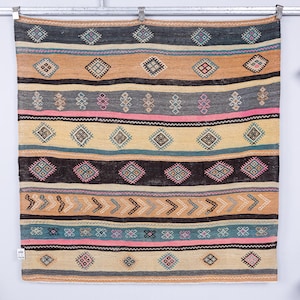 May include: A rectangular, woven rug with a striped pattern. The rug features alternating bands of colour, including tan, grey, black, and pink. Geometric diamond and arrow designs are incorporated throughout the design. The rug is hanging on a metal rod.