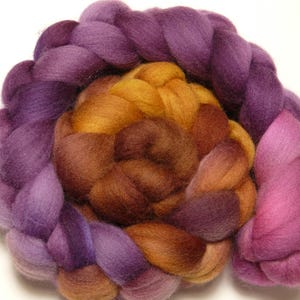 Könnte beinhalten: Nahaufnahme eines Strangs handgefärbter Merinowolle. Die Wolle ist spiralförmig angeordnet und zeigt einen Farbverlauf von tiefem Lila bis zu Goldbraun und Rosa. Die Textur der Wolle ist weich und flauschig.