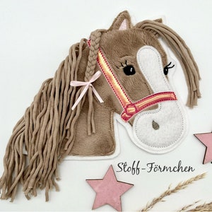 Könnte beinhalten: Ein brauner Plüschpferdekopf mit einem rosa Band und einem rot-orangenen Zaumzeug. Das Pferd hat ein weißes Gesicht mit gestickten Augen und einer rosa Nase. Die Mähne des Pferdes besteht aus braunem Garn.