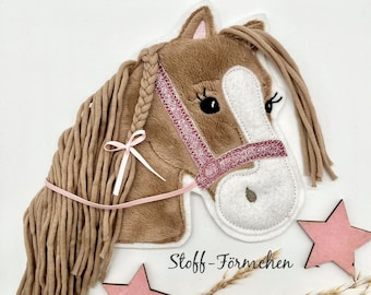 May include: A brown plush horse head with a pink glitter bridle and a white ribbon tied in its mane. The horse head is mounted on a white background with pink stars and dried grass.