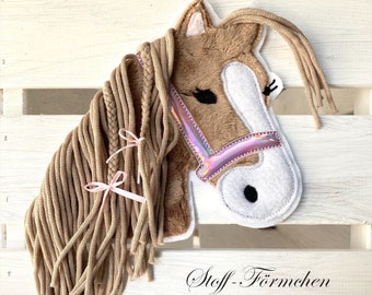 May include: A brown plush horse head with a pink and iridescent bridle. The horse has a long brown mane made of yarn with two pink bows tied in it. The horse head is on a white wooden background with the text "Stoff-Fömchen" in black.