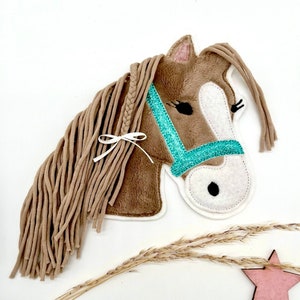 Può includere: Una testa di cavallo di peluche marrone con una briglia turchese e bianca. Il cavallo ha una lunga criniera marrone con un nastro bianco legato in cima.