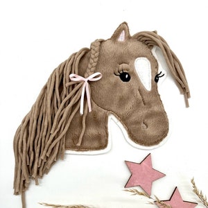 Könnte beinhalten: Ein brauner Plüschpferdekopf mit einem weißen Fleck im Gesicht. Das Pferd hat eine geflochtene Mähne mit einem rosa Band. Der Pferdekopf befindet sich auf einem weißen Hintergrund mit rosa Sternen und trockenem Gras.