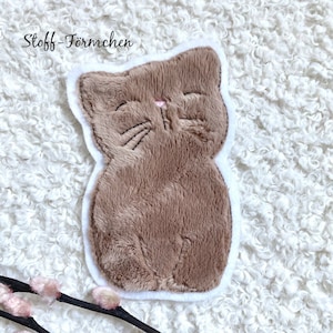 Könnte beinhalten: Ein brauner Plüschkatze mit weißen Nähten an den Rändern. Die Katze hat eine rosa Nase und schwarze gestickte Schnurrhaare. Die Katze sitzt auf einer weißen flauschigen Oberfläche.