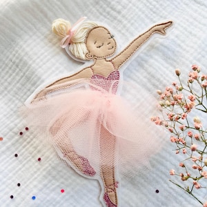 Könnte beinhalten: Stoffapplikation einer Ballerina in einem rosa Tutu. Die Ballerina hat blondes Haar in einem Dutt mit einer rosa Schleife. Ihr Oberteil ist glitzernd rosa und ihr Tutu besteht aus durchsichtigem rosa Stoff. Sie posiert mit einem erhobenen Arm.