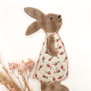 Può includere: Un coniglietto di peluche marrone con un vestito con un motivo floreale di rose rosse su sfondo bianco. Il coniglietto è in piedi, con le orecchie alzate. L'abito ha un bordo rosso. Lo sfondo è bianco, con fiori secchi.