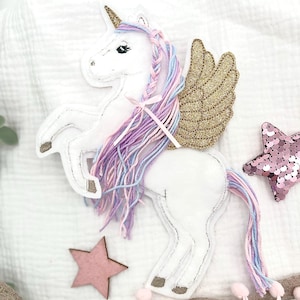 Può includere: Un unicorno di feltro bianco con un'ala di glitter dorato, una criniera e una coda di filato rosa e viola, e un fiocco rosa. L'unicorno è su uno sfondo di tessuto bianco con una stella di legno rosa e una stella di glitter rosa e oro.