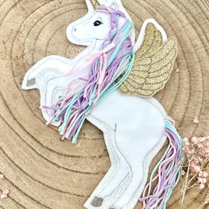 Può includere: Un unicorno di feltro bianco con corno e ali dorate. L'unicorno ha una criniera e una coda multicolore nei toni del rosa, viola e verde acqua. L'unicorno di feltro è su una superficie di legno.