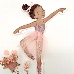 Könnte beinhalten: Stoffapplikation einer Ballerina in einem rosa Tutu, mit glitzerndem Oberteil und Ballettschuhen. Die Tänzerin hat braunes Haar in einem Dutt mit einer rosa Schleife. Die Applikation befindet sich auf weißem Hintergrund mit rosa Dekorationselementen.