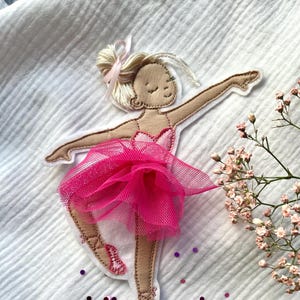 Può includere: Un'applicazione ricamata di ballerina rosa e marrone con una gonna di tulle rosa. L'applicazione è su uno sfondo di tessuto bianco.