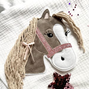 Könnte beinhalten: Ein Pferdekopf aus Filz mit geflochtener Mähne und einem rosa Glitzerhalfter. Das Pferd hat einen braunen Kopf, ein weißes Gesicht und schwarze Augen. Ein rosa Paillettenstern befindet sich neben dem Pferdekopf. Der Pferdekopf ist etwa 20 cm lang.