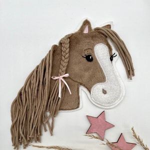 Könnte beinhalten: Ein brauner Plüschpferdekopf mit weißem Gesicht und einem rosa Band in seiner geflochtenen Mähne. Der Pferdekopf ist von getrocknetem Gras und zwei rosa Holzsternen umgeben.