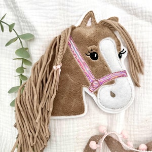 Könnte beinhalten: Ein brauner Pferdekopf mit einem rosafarbenen und schillernden Halfter, braunen Fransenmähne und -schwanz und weißem Gesicht. Der Pferdekopf ist eine Filzapplikation.