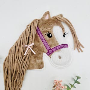 Könnte beinhalten: Ein Plüschpferdekopf mit brauner Mähne und einer rosa Schleife. Das Pferd hat ein weißes Gesicht mit schwarzen Wimpern und einen lila glitzernden Zaum. Das Objekt ist dekorativ.