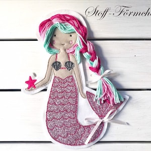 Op de afbeelding: Een vilt mermaid applique met roze en groen garen haar, een roze schelp bh, een roze glinsterende staart en een roze zeester in haar hand. De applique is op een witte houten achtergrond.