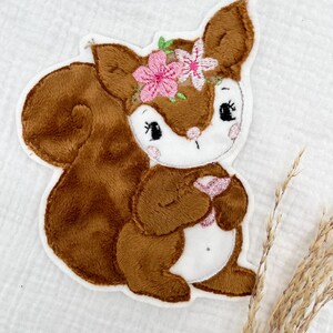 Könnte beinhalten: Gestickter Aufnäher eines Cartoon-Eichhörnchens mit braunem Fell, weißem Gesicht und einem rosa Blumenkranz. Das Eichhörnchen hält ein rosa Herz. Der Aufnäher hat einen weißen Rand.