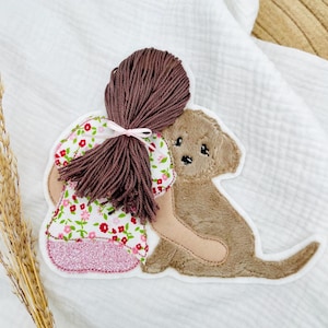 Könnte beinhalten: Ein gestickter Aufnäher mit einem Mädchen, das einen Hund umarmt. Das Mädchen hat braunes Garnhaar, das mit einer rosa Schleife gebunden ist, und trägt ein Blumenkleid mit einem rosa Glitzerrock. Der Hund ist hellbraun.