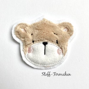 Può includere: Applicazione a forma di orsetto con bordo bianco. L'orso ha una testa di pelliccia marrone, un viso bianco e guance rosa. Il testo "Stoff-Förmchen" è scritto in nero sotto.