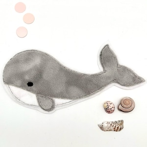 Può includere: Un giocattolo di peluche di balena grigia con pancia bianca e occhi neri. La balena è circondata da cerchi rosa e conchiglie.