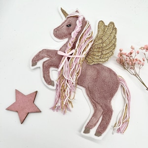 May include: A plush unicorn with a pink body, gold glitter wings, and a pink and gold mane and tail. A pink star is next to the unicorn. The unicorn is standing on its hind legs.