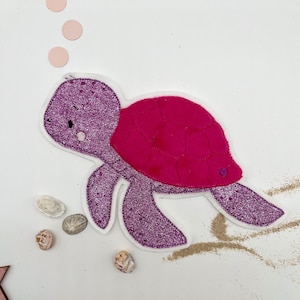 May include: A glittery purple turtle with a bright pink shell, white trim, and small black eyes. The turtle is surrounded by small seashells and pink circles, suggesting an underwater theme. The design is on a white background.