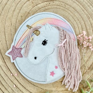 May include: A white felt unicorn with a pink mane and tail, a gold horn, and a rainbow. A pink glitter star is on the left side. The unicorn is on a light gray background.
