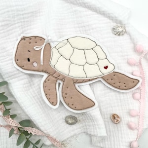 Könnte beinhalten: Eine braune und weiße Filz-Schildkröten-Applikation mit einer weißen Schleife und einem roten Herzen auf ihrem Panzer. Die Schildkröte befindet sich auf einem weißen Stoffhintergrund.