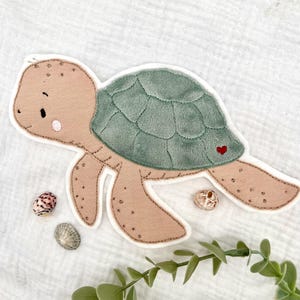 Könnte beinhalten: Ein Applikation einer Schildkröte aus beigem und salbeigrünem Plüsch, mit einem roten Herz auf dem Panzer. Die Applikation hat einen weißen Rand und gestickte Details. Muscheln und Grünzeug sind um die Schildkröte angeordnet.